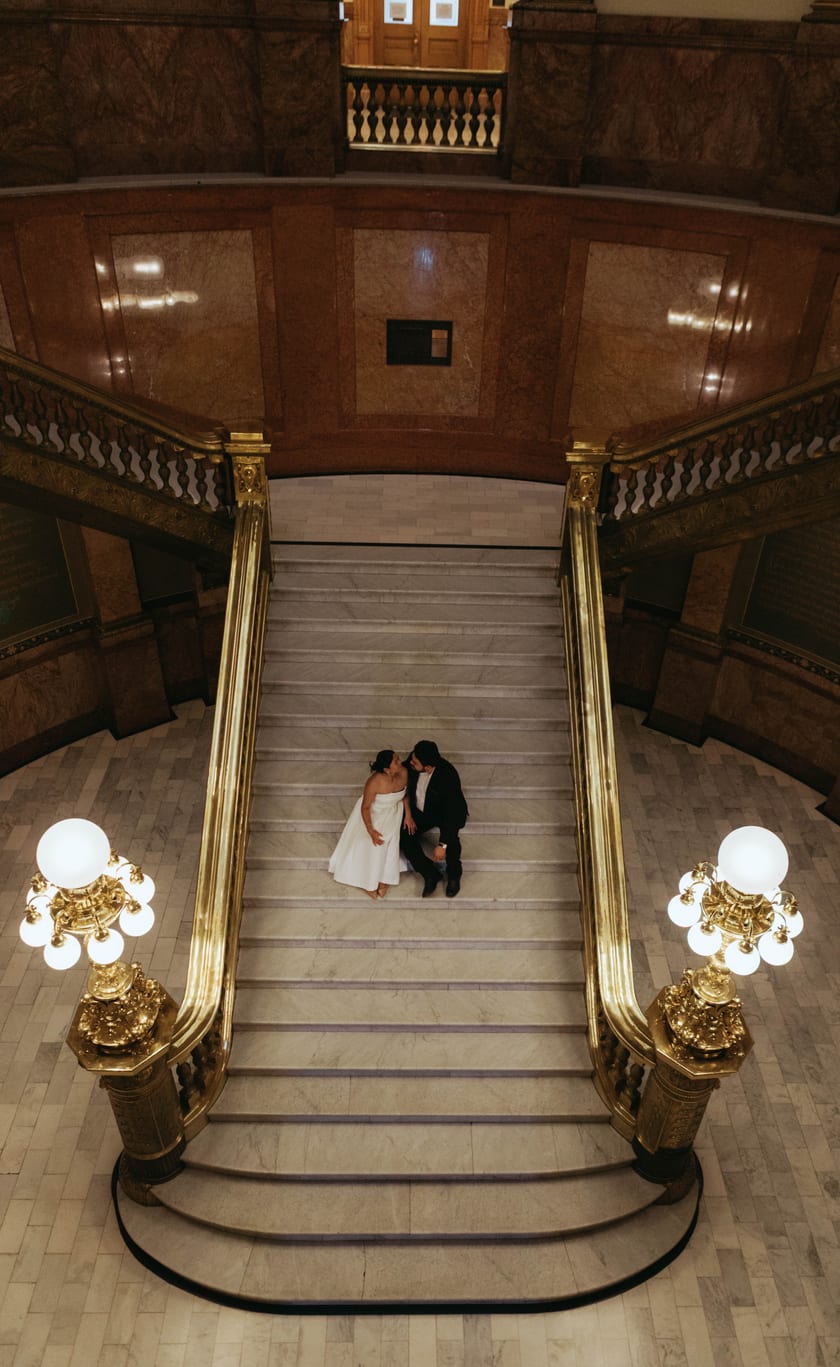 From above: the grand staircase, glowing chandeliers, and the two of them at its foot